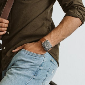 以下が含まれることがあります： 茶色の革製の腕時計バンドと銀色の文字盤の腕時計をしている人。腕時計は左腕につけています。緑のシャツと青いジーンズを着用しています。