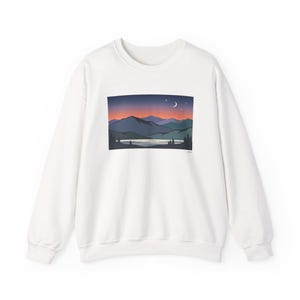 Puede incluir: Una sudadera blanca de manga larga con cuello redondo y un estampado gráfico rectangular en el pecho. El diseño muestra un paisaje con montañas azules superpuestas, un lago tranquilo, pinos en silueta y un cielo al atardecer.
