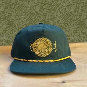 May include: A dark teal cap with a yellow and black rope brim. The cap features a yellow embroidered design of a cross-section of a tree trunk. The background is a light green pattern.