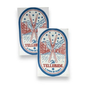 May include: Two white oval stickers with blue borders. Each sticker features a red and white illustration of crossed skis in front of a town with mountains in the background. The text "Ski Telluride" is written in red and white with a snowflake below. The text "A Town of Plenty" is written in red and white below the snowflake.