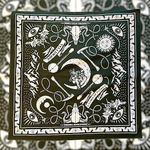May include: Black square bandana with white illustrations of a cowboy playing a banjo on a crescent moon, banjo, pickaxe, sun, eye, and text that reads "Telluride" and "A Town of Plenty". The bandana has a white border and the text "Needle Rock Threads".