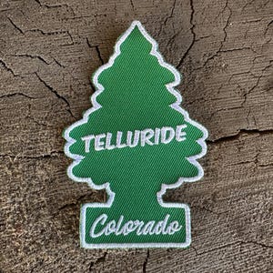 Puede incluir: Un parche bordado verde y blanco con forma de árbol de Navidad estilizado. El parche presenta la palabra "TELLURIDE" en blanco, arqueada en la parte superior, y "Colorado" en blanco en la parte inferior. El parche está sobre una superficie de madera.