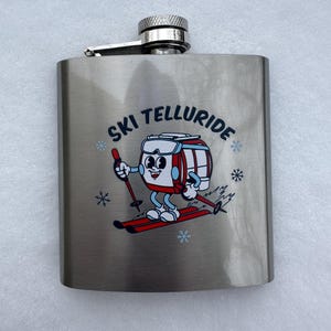 May include: A silver stainless steel flask with a black and red graphic of a cartoon ski lift character skiing down a mountain. The flask has the text "Ski Telluride" printed on it.