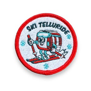 May include: A red, white, and blue embroidered patch with the text "Ski Telluride" and a cartoon ski lift operator skiing down a mountain.