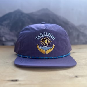 May include: A purple cap with the word "Telluride" embroidered in white above a sun and mountain design. The cap has a blue and white rope detail along the brim and a matching purple brim.