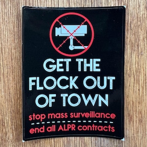 Puede incluir: Una pegatina rectangular negra con un círculo rojo y una línea diagonal sobre un icono de cámara de seguridad gris. El texto dice: GET THE FLOCK OUT OF TOWN, stop mass surveillance, end all ALPR contracts en blanco y rojo.