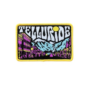 May include: A yellow embroidered patch with the text "Telluride" in a psychedelic font. The patch features a mountain scene with a town in the foreground and a yellow sun in the background.