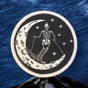 May include: Round black and white patch featuring a skeleton skiing down a crescent moon. The skeleton wears a hat and skis with poles, surrounded by stars. The patch has a white border.