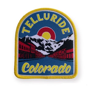Könnte beinhalten: Gelber und blauer gestickter Aufnäher mit dem Text "Telluride Colorado" und einer Darstellung einer Bergstadt mit einer roten und gelben Sonne.