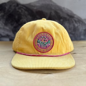 Könnte beinhalten: Eine gelbe Baseballkappe aus Cord mit rosafarbenem Schirm und einem Aufnäher mit der Aufschrift "Telluride, A Town of Plenty".