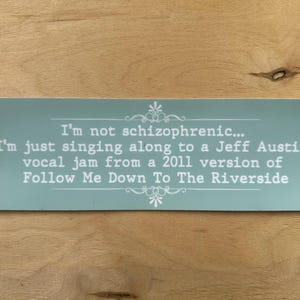 May include: A rectangular, pale turquoise sign with white text and decorative flourishes. The text reads: "I'm not schizophrenic... I'm just singing along to a Jeff Austin vocal jam from a 2011 version of Follow Me Down To The Riverside."