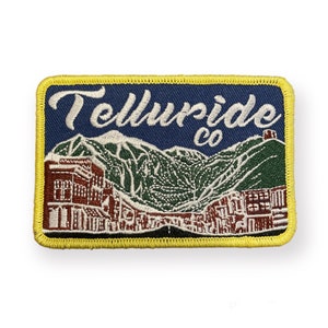May include: Embroidered patch with the text "Telluride co" above a mountain scene and buildings. The patch has a navy blue background with white, green, and brown details, and a yellow border.