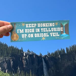 May include: A teal bumper sticker with the text "KEEP HONKING I'M HIGH IN TELLURIDE UP ON BRIDAL VEIL" and a cartoon of a mandolin and a waterfall. The sticker is held up against a backdrop of a mountain and waterfall.
