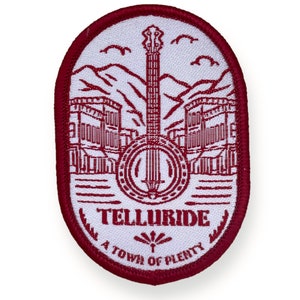 Può includere: Una toppa ricamata rossa con sfondo bianco che presenta un'immagine stilizzata di una città con un banjo al centro. Il testo "Telluride" e "A Town of Plenty" è scritto in rosso sotto l'immagine.