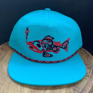 May include: A turquoise blue baseball cap with a black and red embroidered salmon design. The cap has a black and red rope trim around the brim.