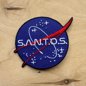 Könnte beinhalten: Ein blauer und roter gestickter Aufnäher mit einer weißen Umrandung eines Raumschiffs und dem Wort "SANTOS" in weißen Buchstaben.