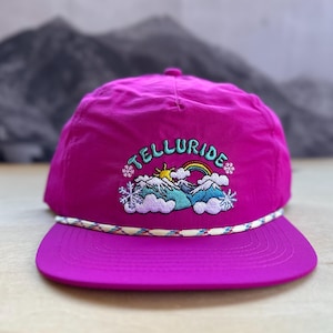 May include: A pink nylon baseball cap with a white rope trim and an embroidered design featuring the word "Telluride" with a rainbow, mountains, and snowflakes.