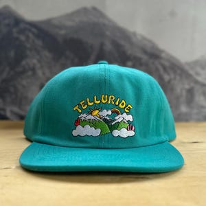 Può includere: Un berretto da baseball blu turchese con un ricamo a forma di catena montuosa, un sole, un arcobaleno e la parola "Telluride".