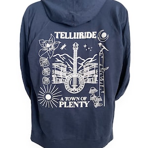 May include: A navy blue hooded sweatshirt with a white graphic design on the back. The design features a banjo, a mountain range, a town, and the text "Telluride, A Town of Plenty".