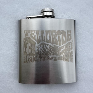 Könnte beinhalten: Eine Edelstahl-Taschenflasche mit Schraubverschluss und Scharnierarm. Die Flasche ist mit dem Wort "TELLURIDE" über einer Bergkette und Gebäuden graviert.