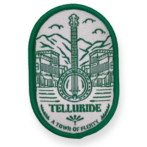 Könnte beinhalten: Ein ovales Abzeichen mit grünem Rand und weißem Innenraum. Das Design zeigt ein Banjo, Berge, Gebäude und den Text "TELLURIDE A TOWN OF PLENTY" in Grün.