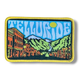 May include: A rectangular embroidered patch with a yellow border featuring a mountain landscape and town buildings in orange and brown. The word TELLURIDE is written in large, light blue letters across the top, with a yellow sun in the sky.