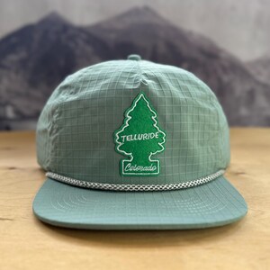 Può includere: Un berretto da baseball in ripstop verde salvia con un cordino intrecciato bianco alla base della corona. Una toppa in feltro verde a forma di pino è centrata sul davanti, con il testo ricamato in bianco Telluride e Colorado.