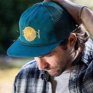 May include: A teal blue corduroy and mesh trucker hat with a yellow embroidered fish design. The fish has a circular pattern inside.