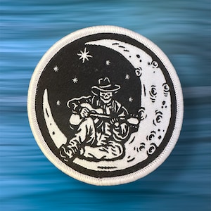 May include: Circular patch with a black and white design of a skeleton playing a guitar on a crescent moon. The patch includes stars and a white border. The design is reminiscent of a folk art style.