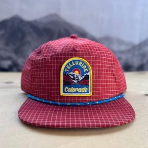 Peut inclure: Casquette de baseball à carreaux rouges et blancs avec un patch bleu et jaune qui dit "Telluride Colorado" et représente une scène de montagne.