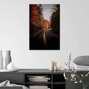 May include: A winding road through a forest with vibrant fall foliage. The road is lined with trees with red, orange, and yellow leaves. The road is paved and has two yellow lines down the center.