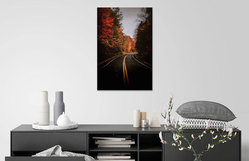 May include: A winding road through a forest with vibrant fall foliage. The road is lined with trees with red, orange, and yellow leaves. The road is paved and has two yellow lines down the center.