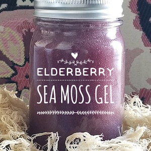 May include: A glass jar filled with a dark purple liquid, labeled "Elderberry Sea Moss Gel". The jar has a silver lid and is sitting on a bed of dried sea moss.
