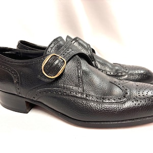 May include: A pair of black leather dress shoes with a textured finish. The shoes feature a gold-colored buckle closure and decorative perforations along the edges. The shoes have a low heel.