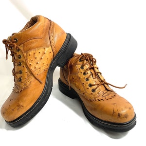 May include: A pair of tan leather boots with black soles. The boots feature a textured design on the upper, lace-up closures, and decorative stitching. The boots are designed for outdoor wear.