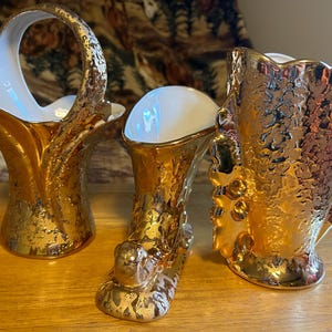 May include: Three gold-plated ceramic vases with a textured surface. The vases are in the shape of a basket, a boot, and a tall, slender vase.