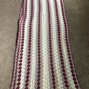 May include: A long, white crocheted table runner with burgundy stripes and a repeating pattern of circles.