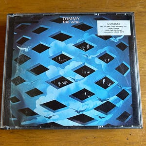 May include: A CD case with a blue and black geometric design featuring a pattern of overlapping squares. The text "TOMMY the WHO" is printed in white on the front of the case. The CD case is a promotional item for "SMG Direct Marketing, Inc."