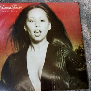 May include: A vintage vinyl record cover featuring a woman with long dark hair wearing a black and white striped jacket. The cover is red and has the text "Grenne Elliman" and "Night Flight" on it.