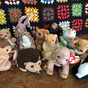 May include: A collection of 11 plush toys, including a brown owl, a brown hedgehog, a brown bear, a brown squirrel, a white rabbit, a brown and white squirrel, a brown and white hamster, a brown and white deer, a green frog, a brown bear, and a black and white skunk. The toys are all sitting on a wooden surface.