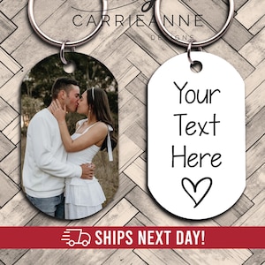 May include: Two metal dog tag keychains with silver split rings. One keychain features a color photograph of a couple kissing outdoors. The other keychain is white with the black printed text Your Text Here and a hand-drawn heart symbol below it.