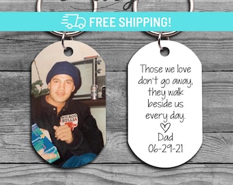 May include: A white oval keychain with the text "Those we love don't go away, they walk beside us every day. Dad 06-29-21". The keychain is attached to a silver key ring. A second oval keychain with a photo of a man wearing a blue hat and a red jacket with the words "Chicago Bulls" on it.