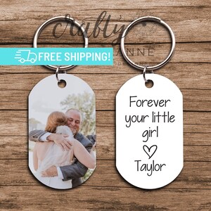 May include: Two oval-shaped keychains. One keychain features a photo of a bride and her father hugging. The other keychain is white with black text that reads "Forever your little girl ♥ Taylor".
