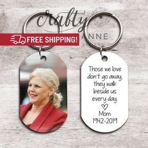 May include: Two keychains with silver rings. One keychain features a photo of a woman in a red jacket. The other keychain has the text: "Those we love don't go away, they walk beside us every day. Mom 1942-2019."