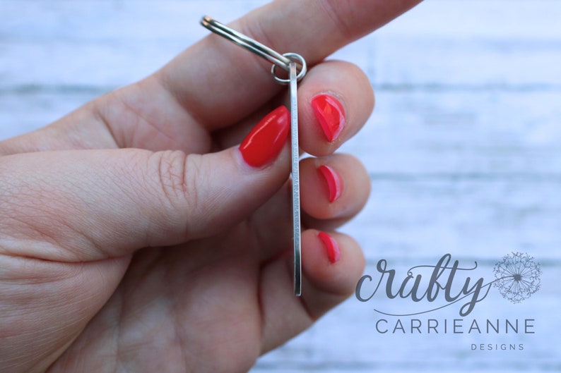 May include: Silver key ring with a rectangular tag. The tag is blank and has a brushed metal finish. The keyring is held in a hand with red nail polish.