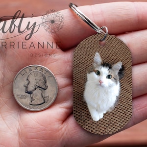 K&ouml;nnte beinhalten: Ein brauner ovaler Schl&uuml;sselanh&auml;nger mit einer wei&szlig;en Katze mit gr&uuml;nen Augen. Der Schl&uuml;sselanh&auml;nger wird in einer Hand gehalten und eine 25-Cent-M&uuml;nze liegt zum Gr&ouml;&szlig;envergleich daneben.