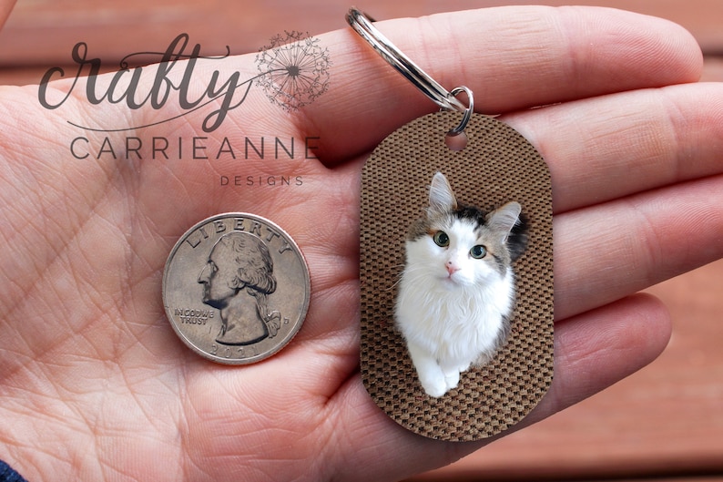 K&ouml;nnte beinhalten: Ein brauner ovaler Schl&uuml;sselanh&auml;nger mit einer wei&szlig;en Katze mit gr&uuml;nen Augen. Der Schl&uuml;sselanh&auml;nger wird in einer Hand gehalten und eine 25-Cent-M&uuml;nze liegt zum Gr&ouml;&szlig;envergleich daneben.