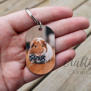 May include: A brown and white guinea pig wearing a black and white bow tie is featured on a metal keychain tag. The tag is oval shaped and has a silver ring.