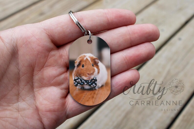 May include: A brown and white guinea pig wearing a black and white bow tie is featured on a metal keychain tag. The tag is oval shaped and has a silver ring.