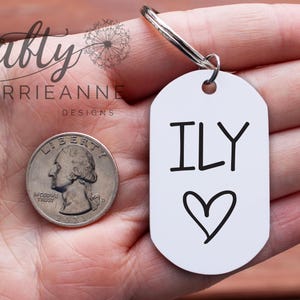 May include: White metal keychain with the text "ILY" and a heart drawn on it. The keychain is being held in a hand.